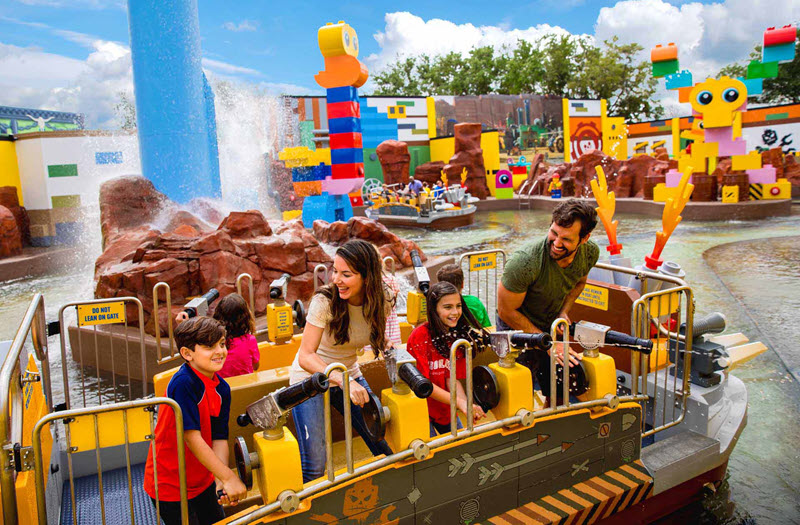 A family rides a water attraction at an amusement park decorated with large colorful LEGO structures, while spraying water with mounted cannons.