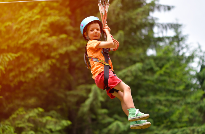 A happy little boy riding a zip line with a forest of trees behind him.