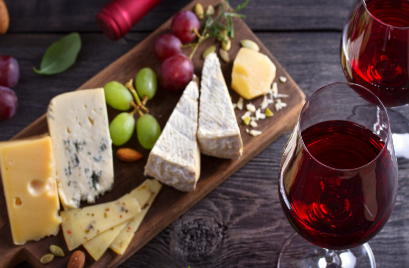 Two glasses with red wine placed next to a platter with various cheeses.