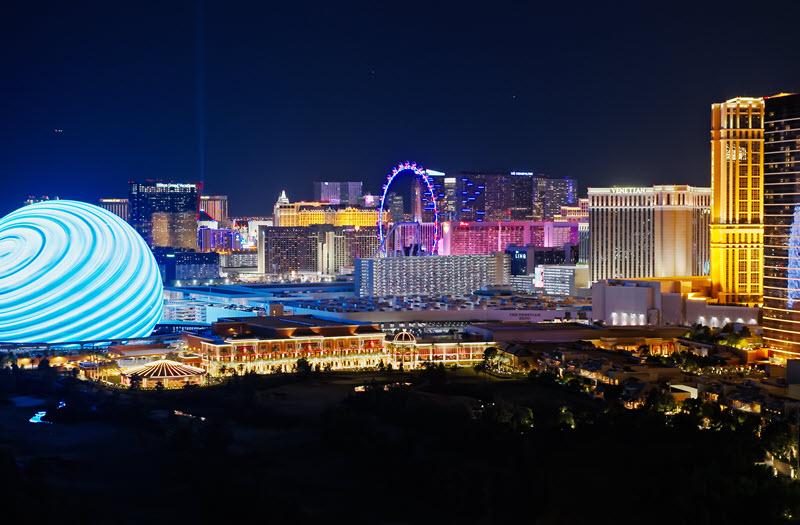 Nighttime view of the Las Vegas Strip with brightly lit hotels, casinos, the illuminated sphere, and a large Ferris wheel in the background.
